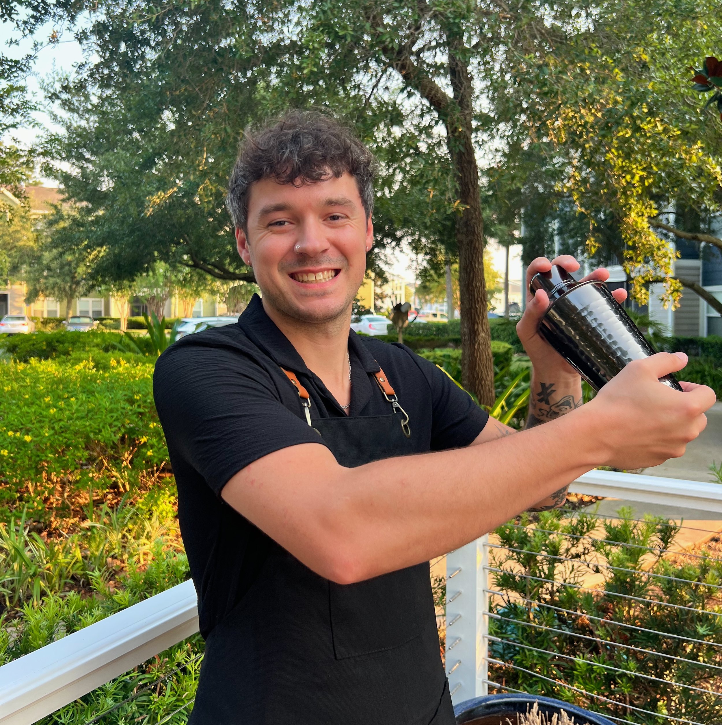 Young man smiling and holding a bartender shaker tin outdoors in a park with trees, bushes in a pretty atmosphere