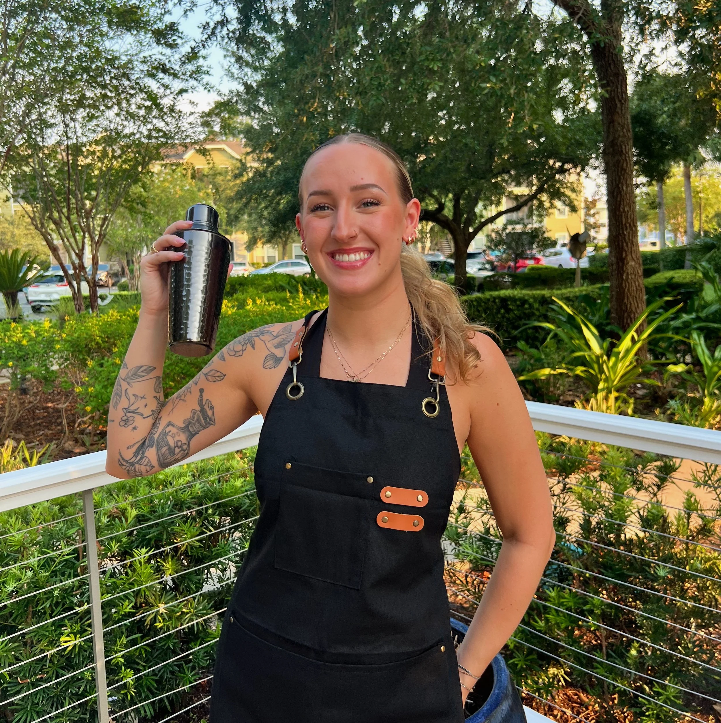 Young woman smiling and holding a bartender shaker tin outdoors in a park with trees, bushes in a pretty atmosphere.