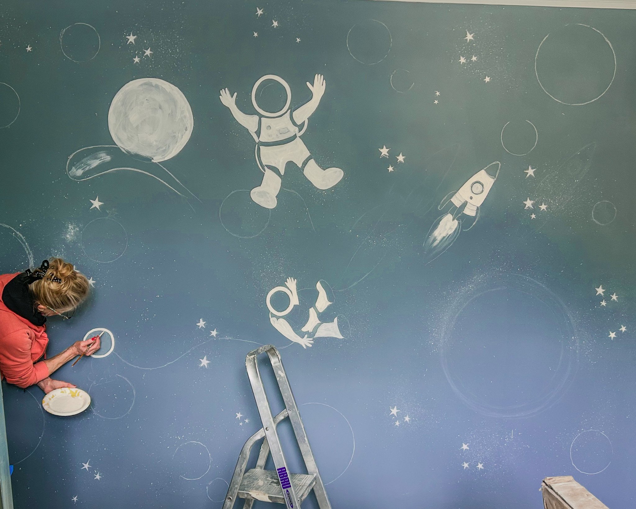 Person painting a space-themed mural with stars, planets, an astronaut, and a rocket on a blue wall.
