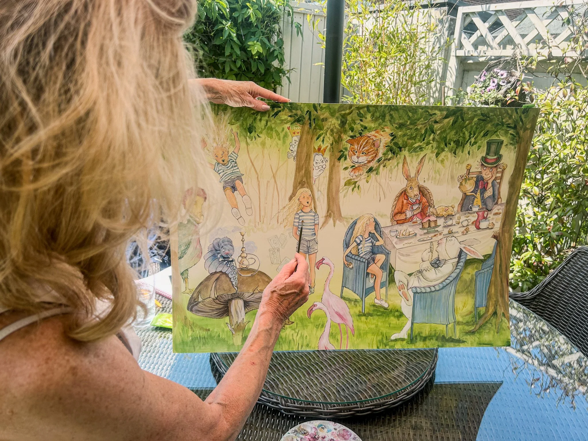 An elderly woman with long curly blonde hair is painting a colorful illustration of animals and characters in a forest setting outdoors. She is holding a brush with her right hand and the painting features various animals and characters including a rabbit, and a person wearing a top hat, among others.