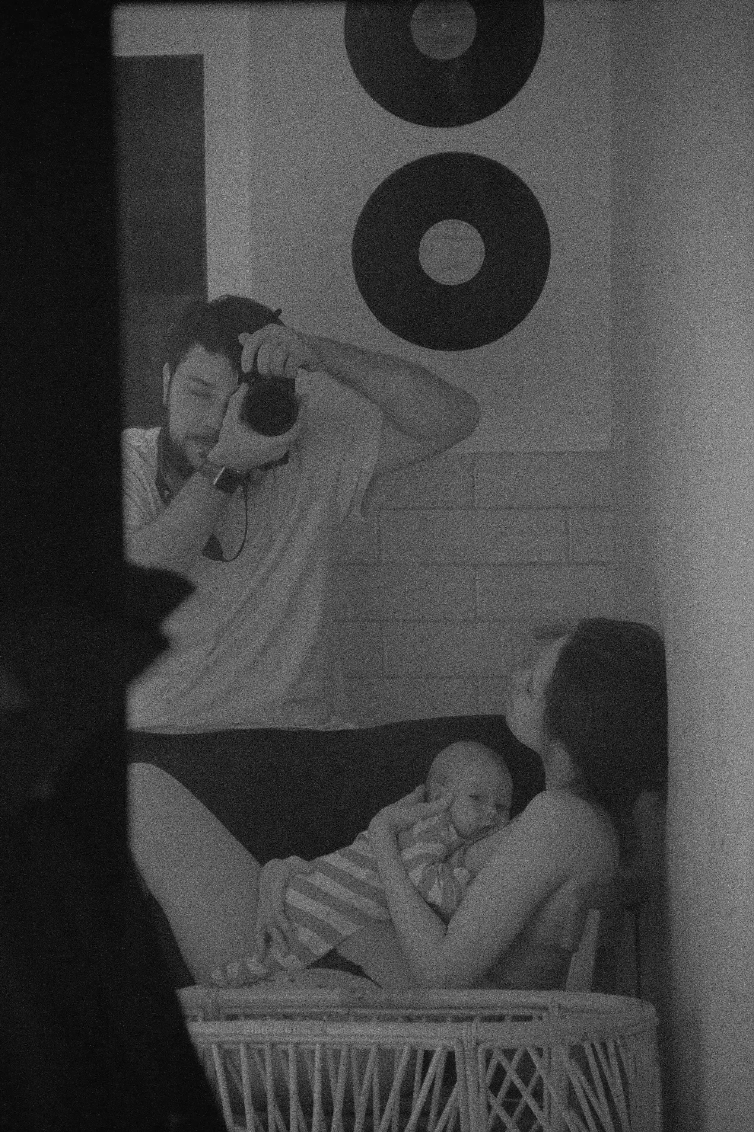 A black and white photo of a newly postpartum mom reclined and breastfeeding her newborn baby. The new dad looks on and takes a photo of the happy family.
