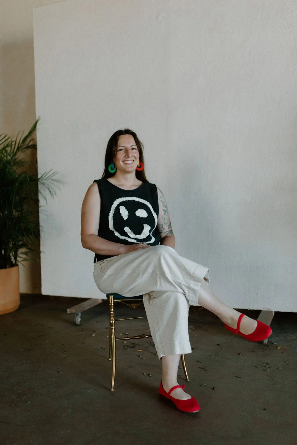 Ale Falco, Minnesota birth doula of La Doula, sits in a chair, legs crossed, hands in lap, & smiling. She has bright red shoes, light comfy pants, a black sweater with a smiley face, & funky earrings. She has white skin & long brown hair.