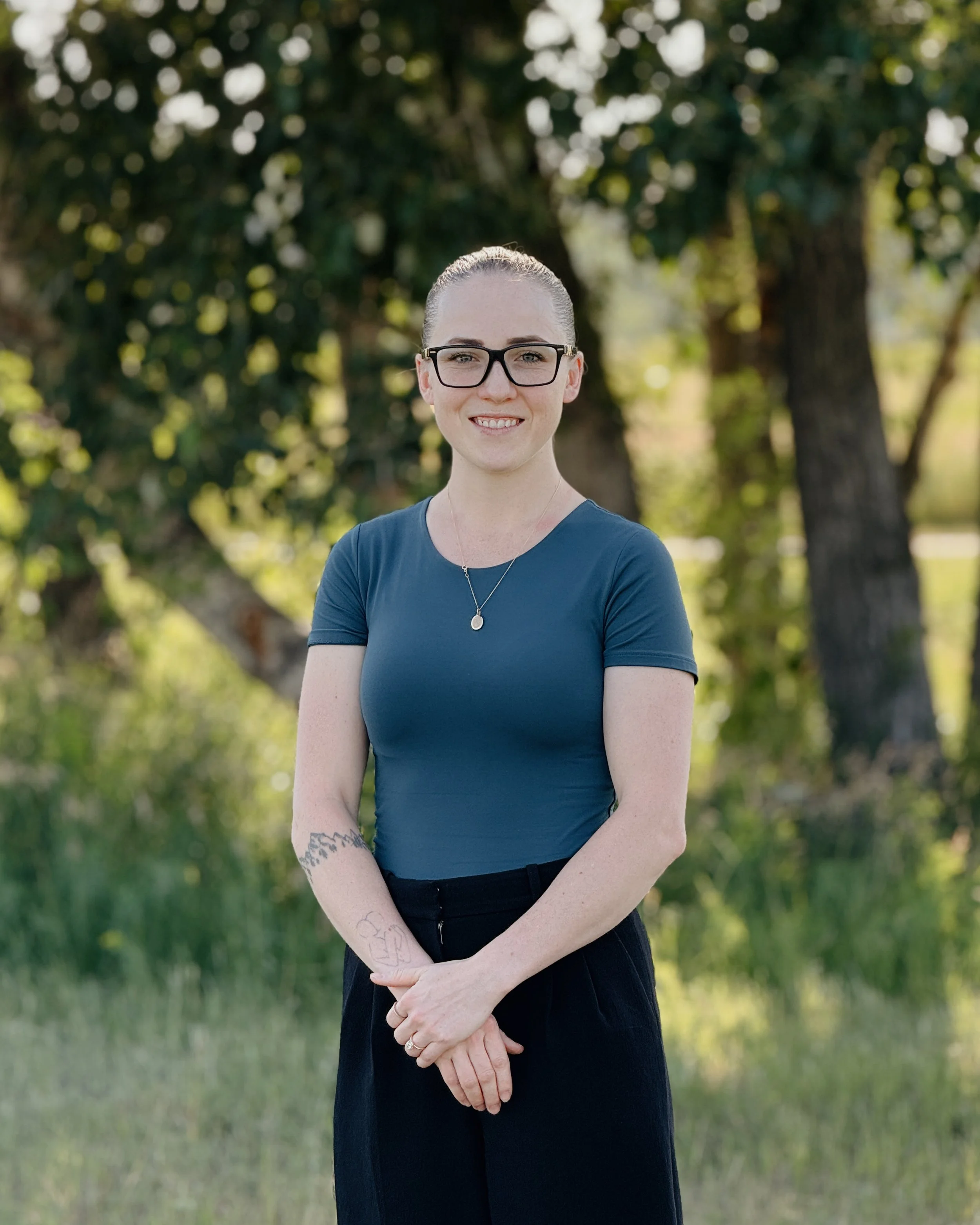 A young woman with glasses and a bun hairstyle standing outdoors in front of a leafy background, wearing a blue shirt and black pants, smiling with her hands crossed in front of her.
