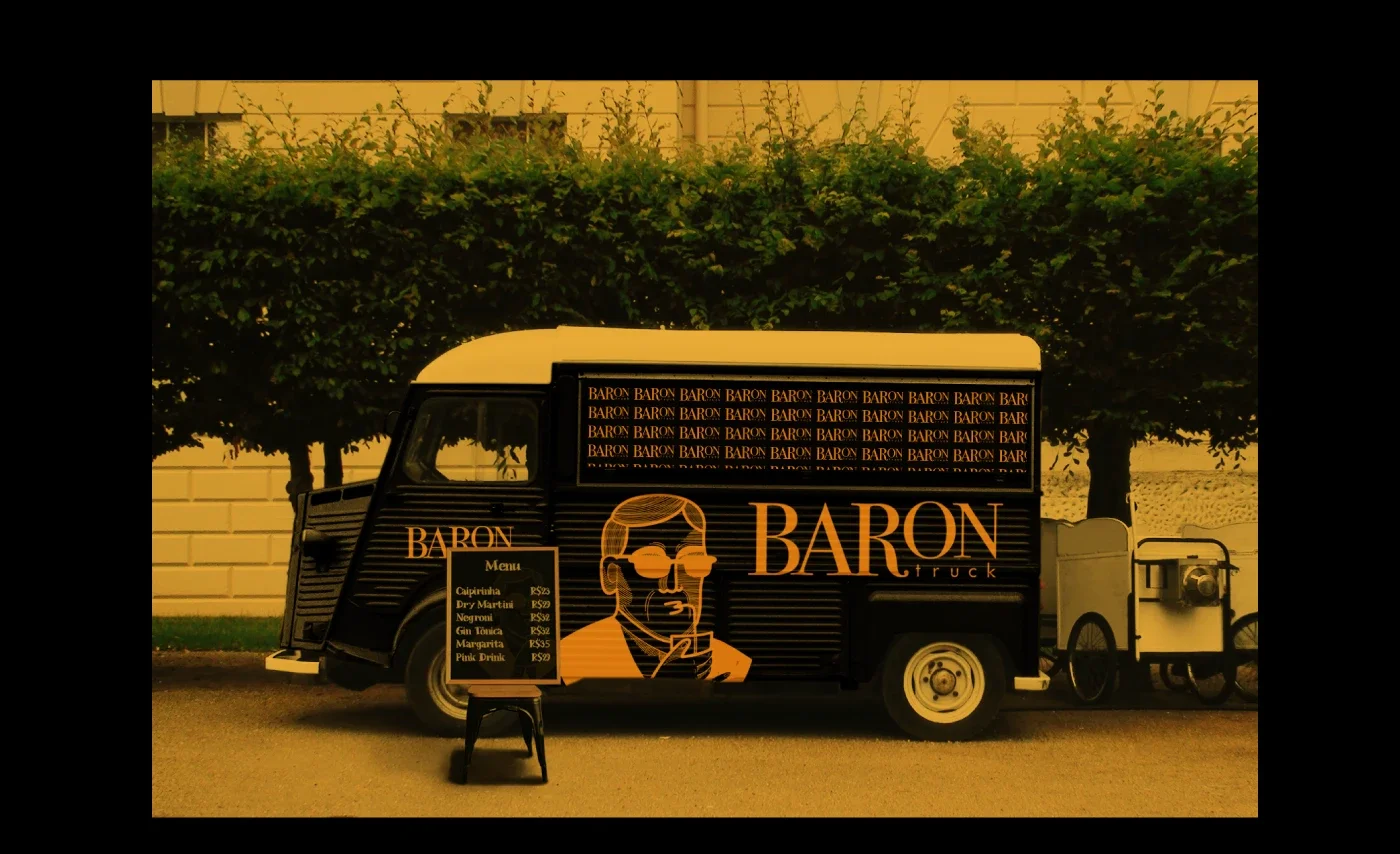 Food truck decorado com o nome 'Baron' e uma ilustração de um homem de óculos. Ao lado, um menu com opções de bebidas e preços. No fundo, árvores e uma parede amarela.