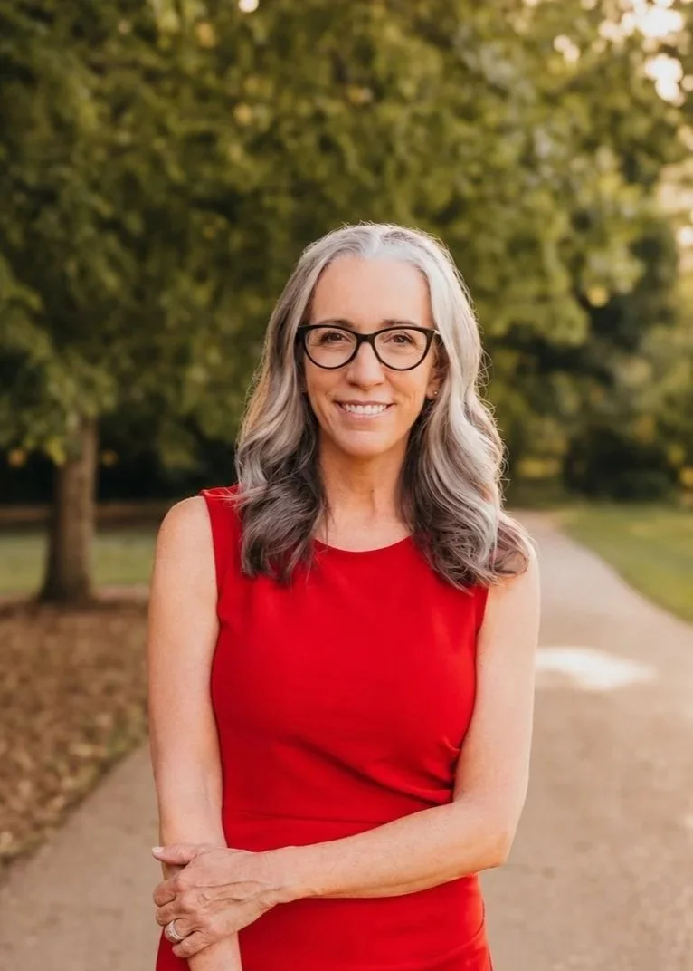 A smiling middle-aged woman with gray hair and glasses wearing a sleeveless red dress standing outdoors on a pathway surrounded by green trees.