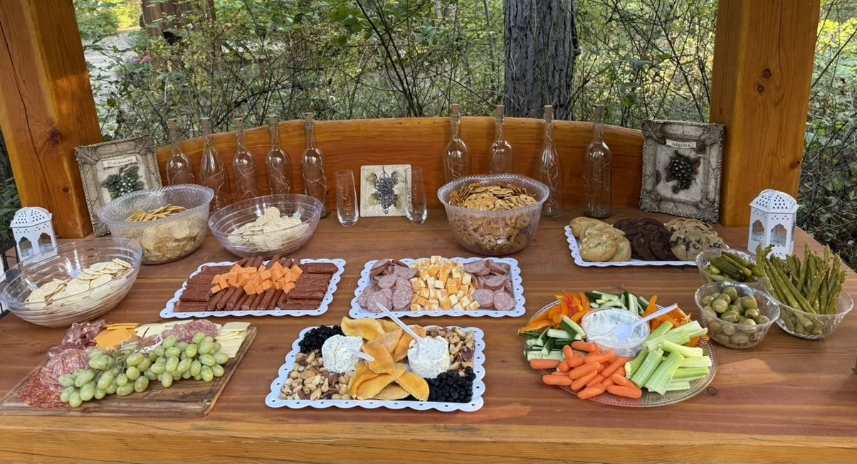 Outdoor picnic table with a variety of snacks including cheese, meats, vegetables, crackers, fruit, dips, and popcorn, set in a wooded area.