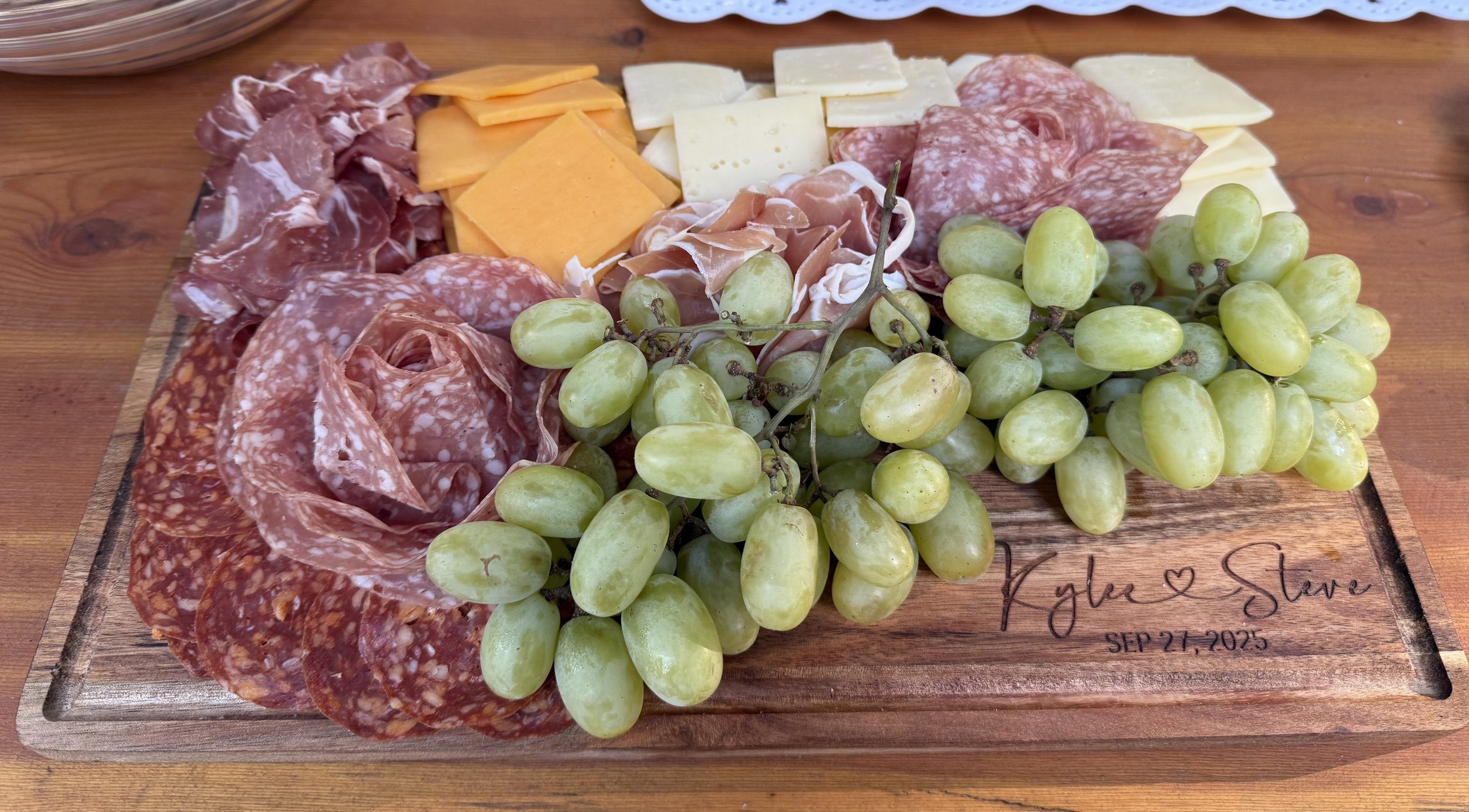 A wooden cheese and charcuterie board with green grapes, sliced cheese, and various cured meats on a wooden table.