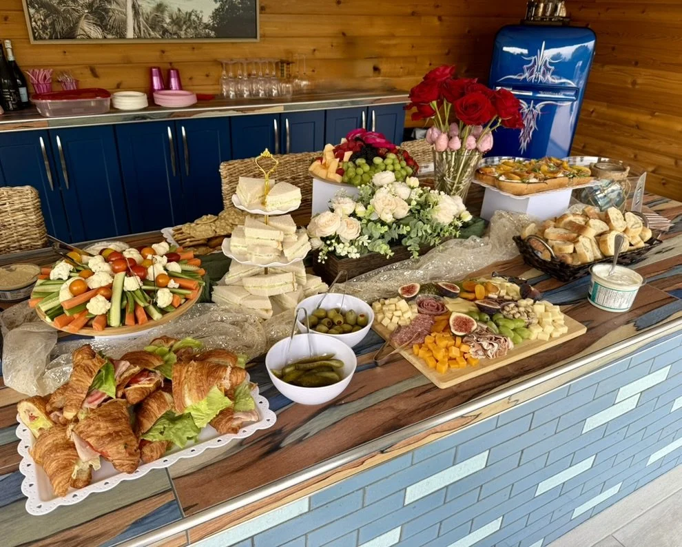 Buffet table with various appetizers, sandwiches, vegetables, cheeses, fruits, and flowers in a cozy indoor setting.