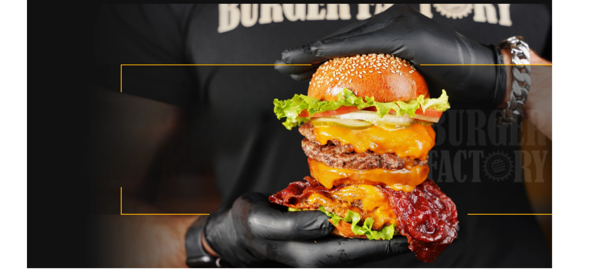 A person holding a large burger with various ingredients, including lettuce, tomato, pickles, cheese, burger patty, bacon, and sesame seed bun, wearing black gloves.