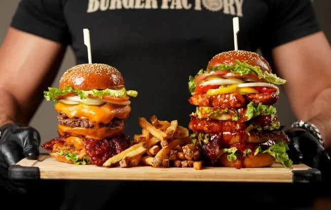 A person holding a wooden tray with two large burgers and fries.
