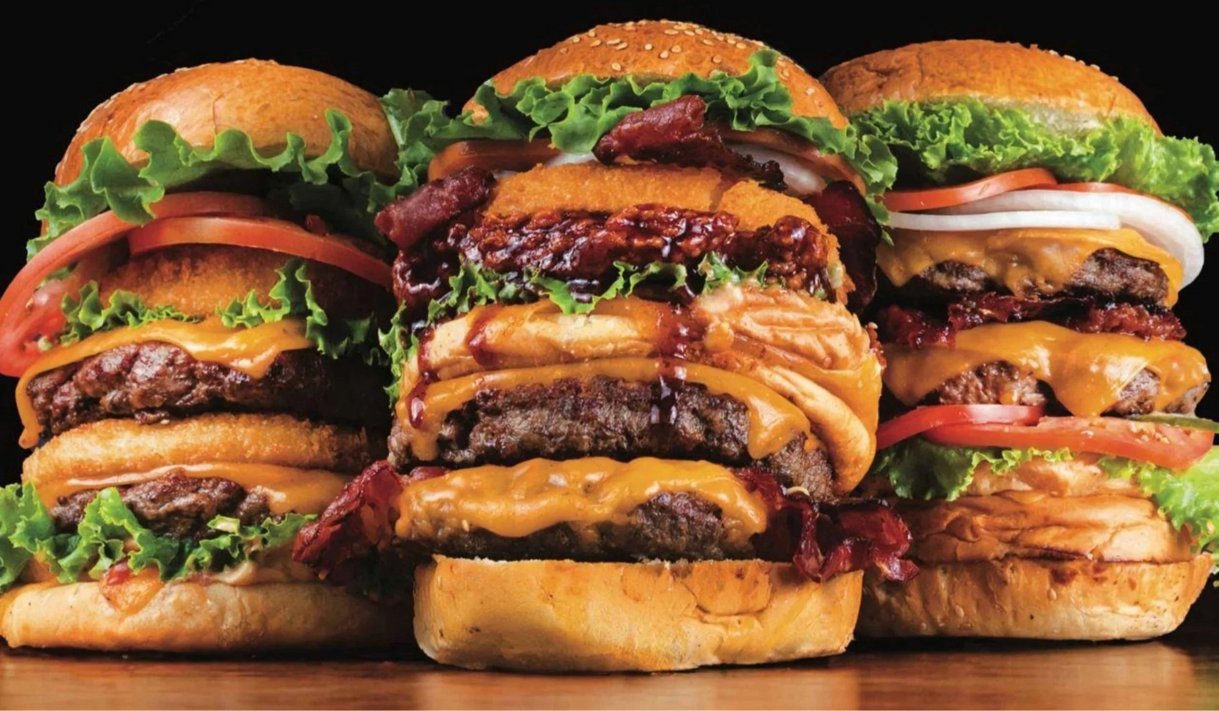 Three loaded cheeseburgers with lettuce, tomato, onions, bacon, and cheese, stacked together on a wooden surface.