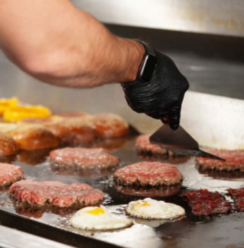 Person cooking burger patties and eggs on a flat-top grill.