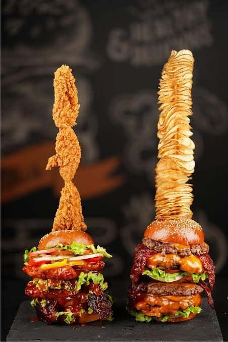 Two tall gourmet burgers with onion rings and fried chicken tenders on top, served with crispy onion rings on skewers, with shredded cheese and lettuce.