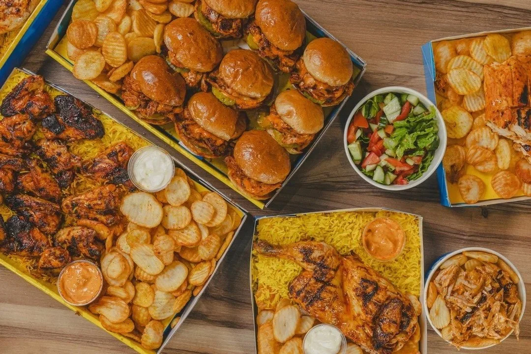 Variety of fast food including mini burgers, chicken wings, potato chips, salad, grilled chicken, and a casserole with onions on a wooden table.