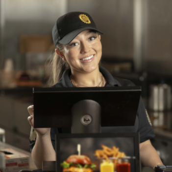 A smiling woman in a uniform and cap standing at a restaurant counter, facing a point-of-sale screen.