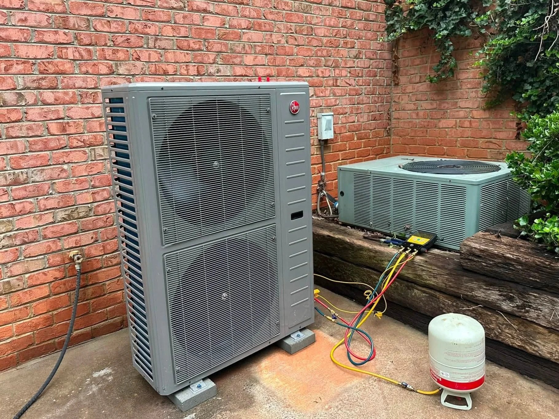 An outdoor view of two air conditioning units installed near a red brick wall with green ivy growing on the wall's corner. The units are connected by colored electrical wires, and one has a Rhoss brand label. There is a white container on the ground 
