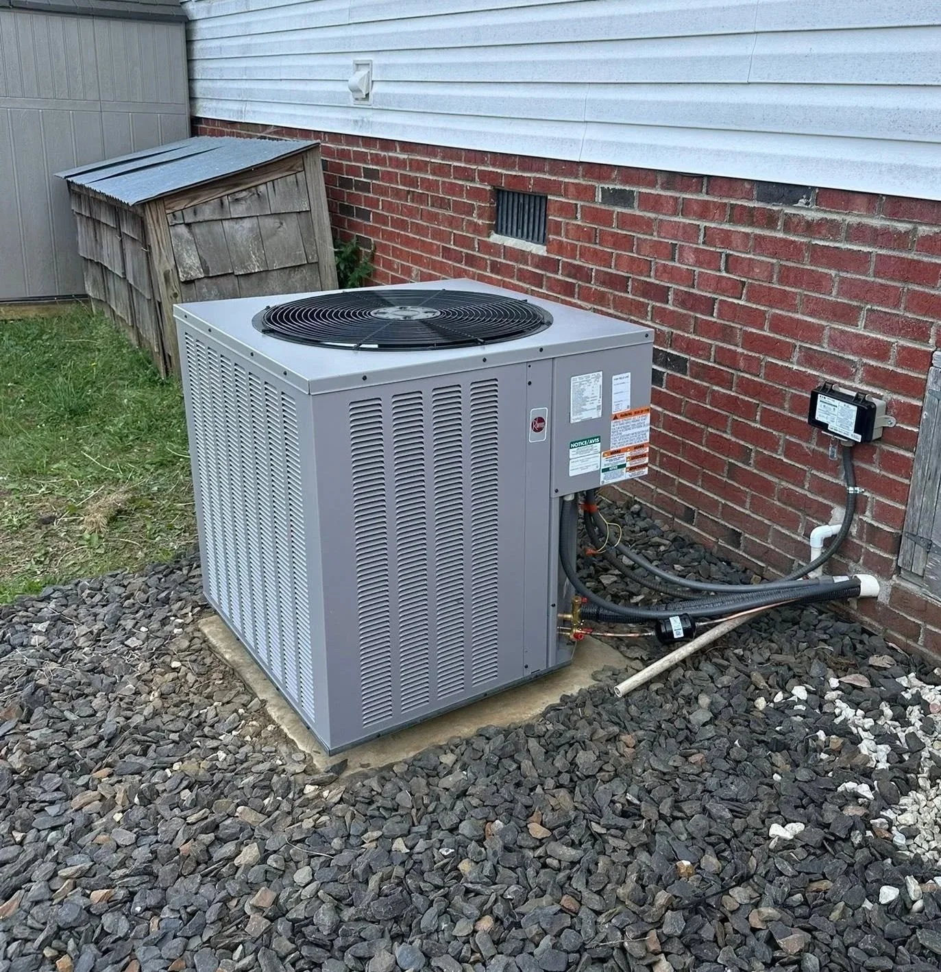 Exterior view of a house showing an air conditioning unit on a small concrete pad, with some rocks and gravel on the ground. The house has white siding and a brick foundation with small windows, a vent, and an electrical box visible.