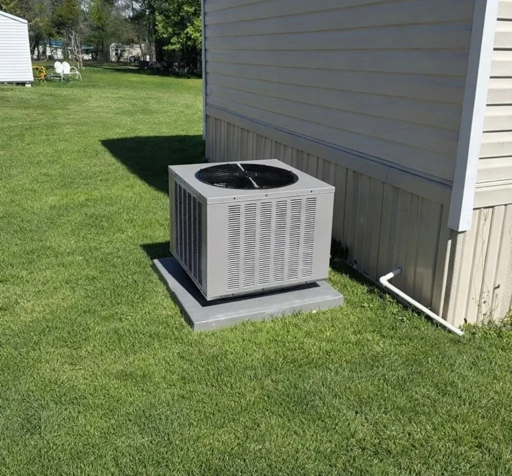 Exterior view of a residential house's side yard with a gray HVAC unit on a concrete pad, next to the house's beige siding, and green lawn extending into the background with trees and other houses.