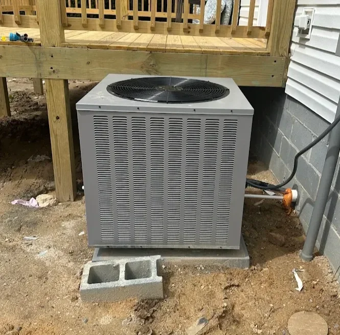 An outdoor air conditioning unit installed on a concrete block beside a house with a deck above. Electrical wiring and meters are visible on the house wall.