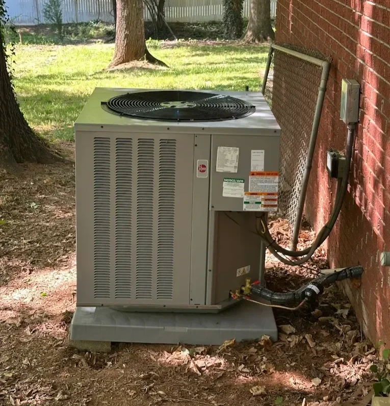 Exterior air conditioning unit with fan on top, placed on a concrete pad near a brick wall, connected to household electrical and refrigerant lines.