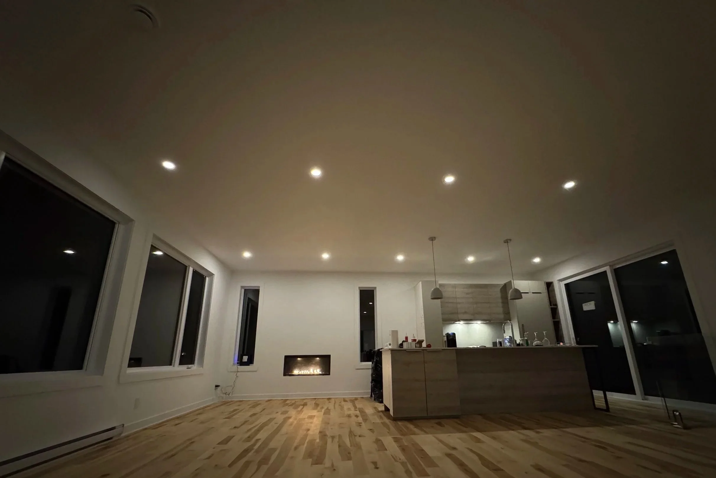 A spacious modern kitchen with a high ceiling, recessed lighting, large windows, and a wooden floor.