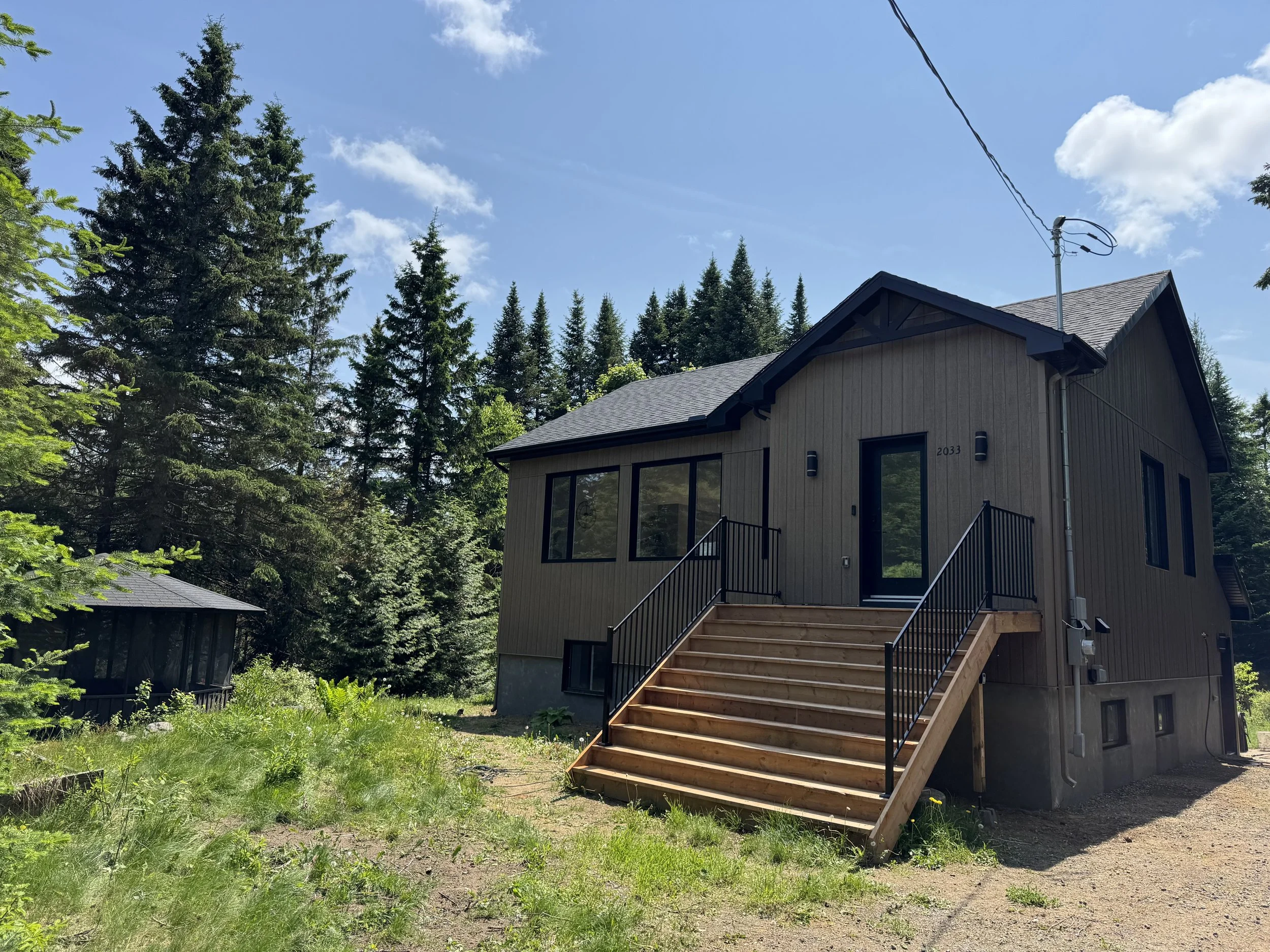 A two-story house with tan siding and a staircase leading to a back door, surrounded by tall evergreen trees and a grassy yard, under a blue sky with some clouds.