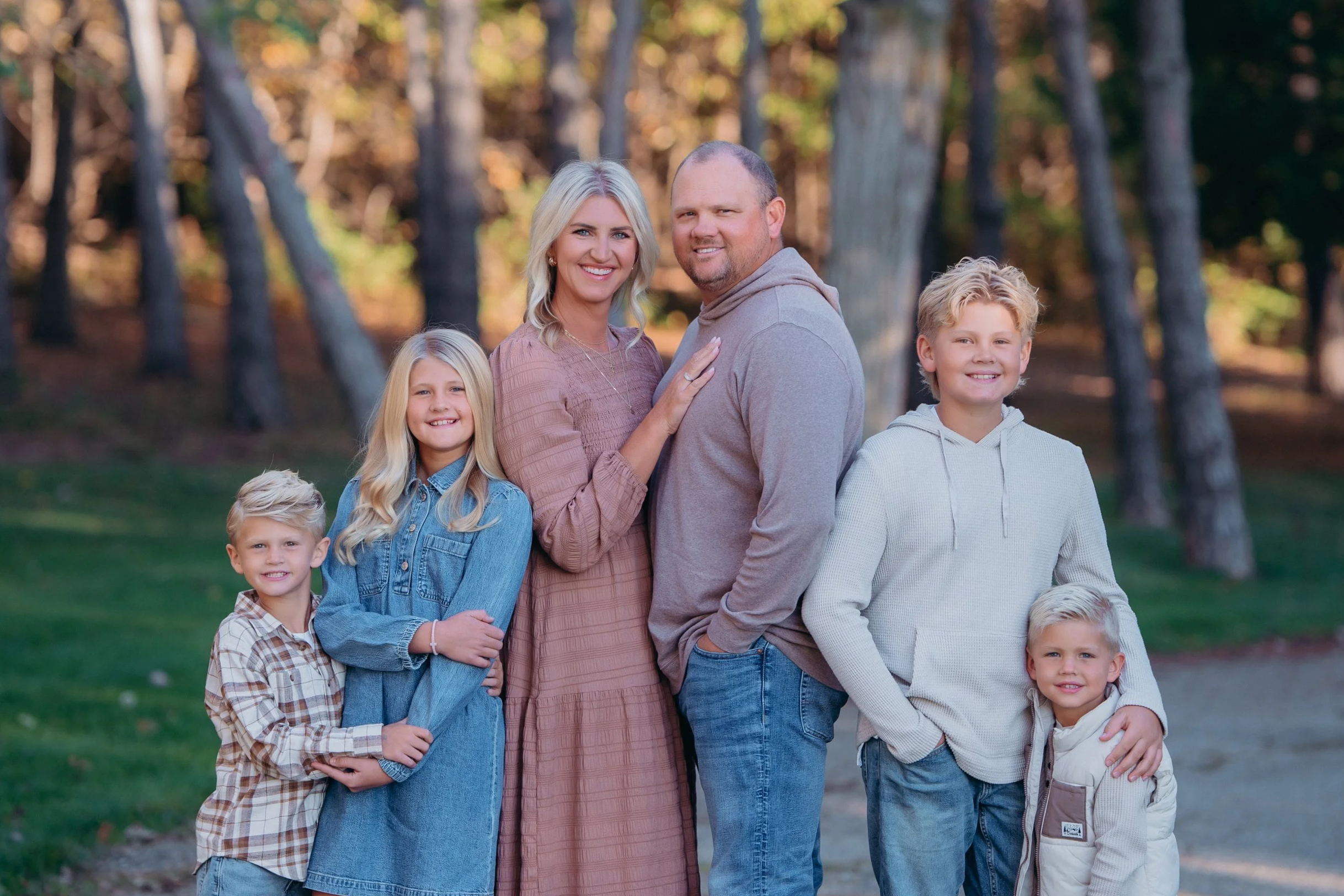 A smiling family of seven posing outdoors in a wooded park during autumn. The mother and father are in the center with three children standing around them, two boys and one girl, all smiling and dressed casually.