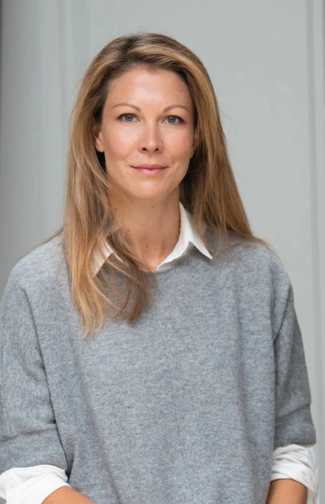 A woman with light brown hair and blue eyes, wearing a white collared shirt under a gray sweater, standing indoors in front of a plain light-colored wall.
