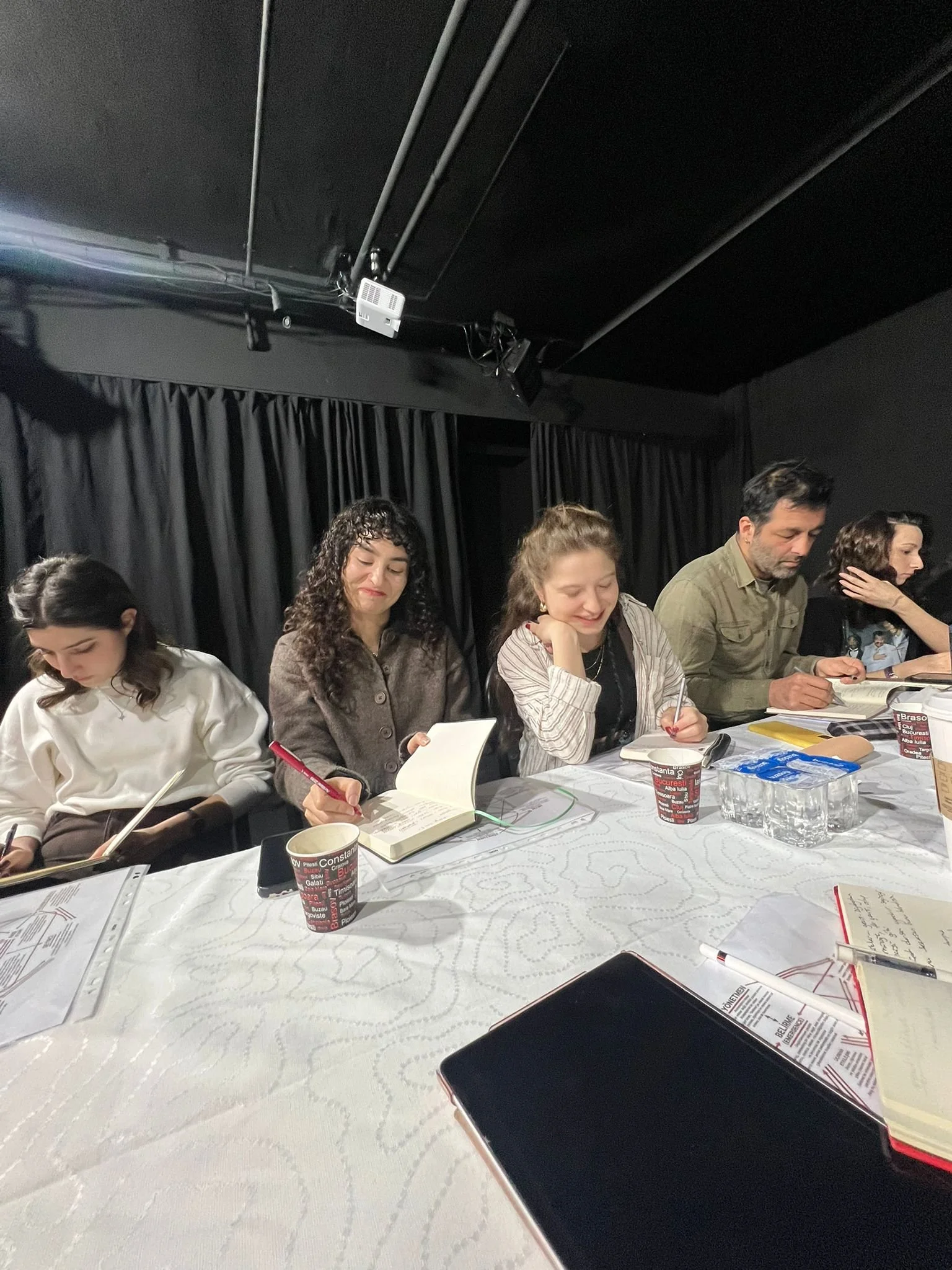 People sitting at a meeting table with notebooks, pens, cups, and papers, in a dark room with black curtains and ceiling.