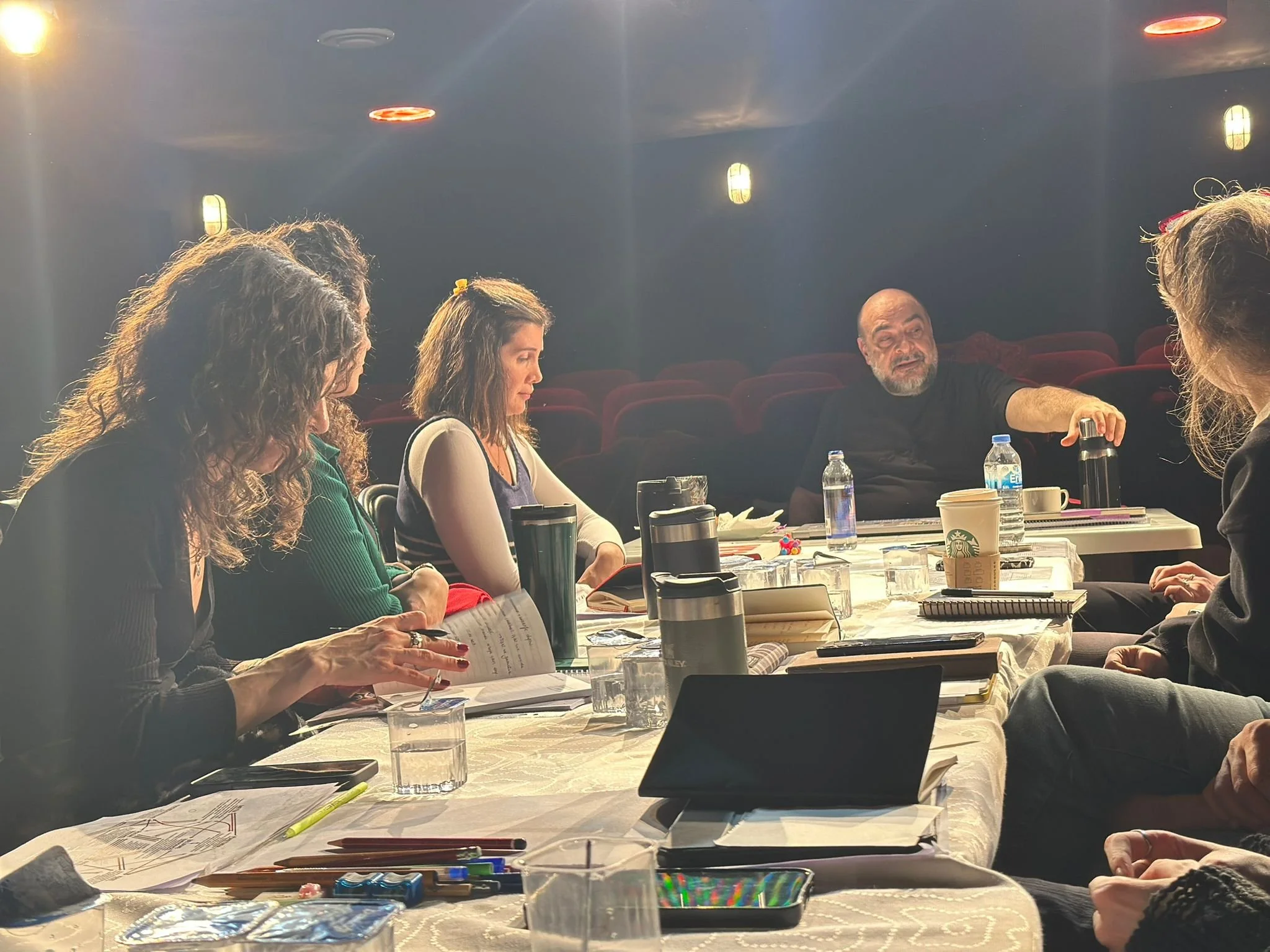 A group of people sitting around a large table in a room with dark walls and red velvet seats in the background. They are engaged in a discussion or meeting, with various papers, notebooks, pens, and beverages on the table. There are multiple glasses