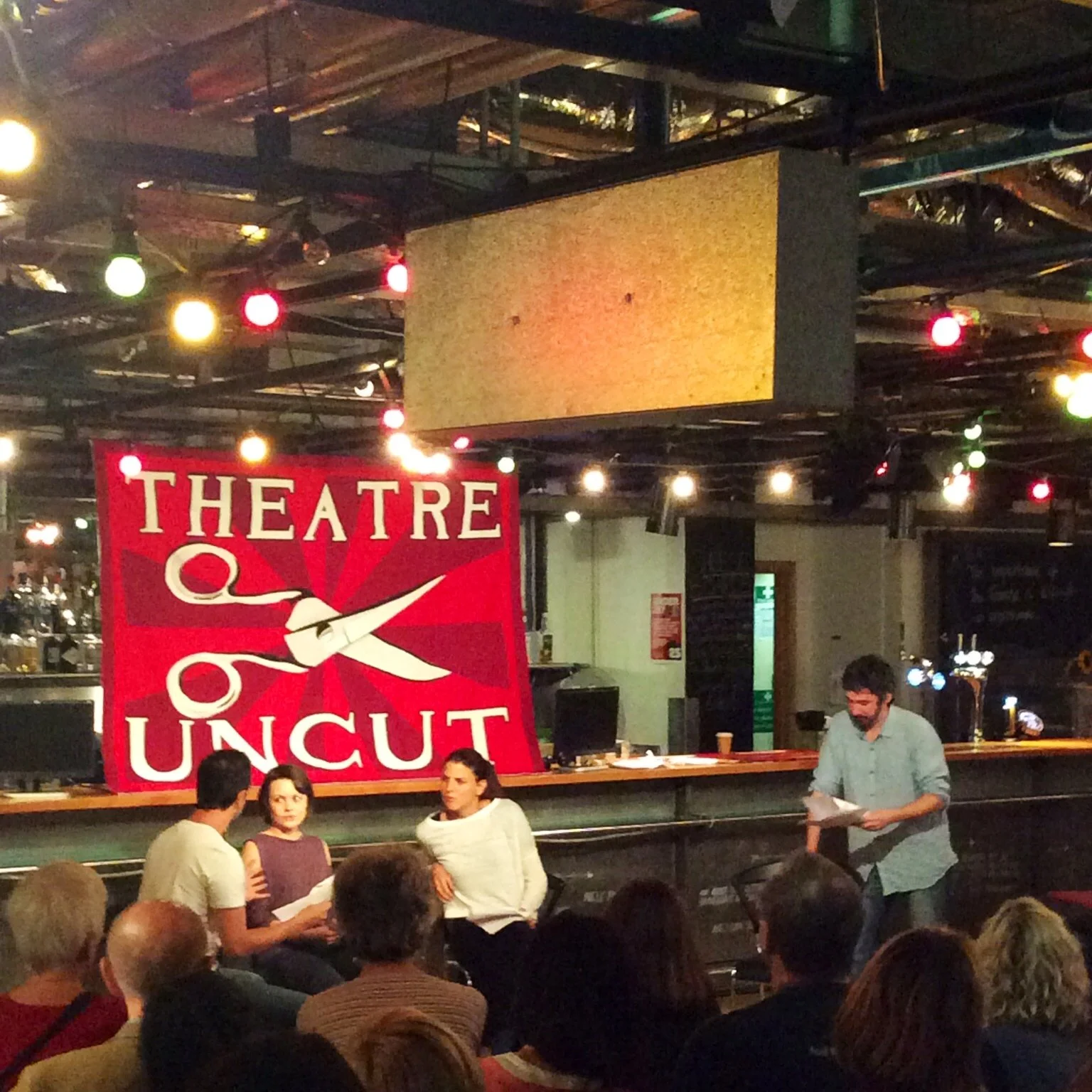 People seated and standing in a dimly lit theater or performance venue, with a large red and white banner reading 'Theatre Uncut' featuring a pair of scissors and red rays in the background. The ceiling has multicolored lights.