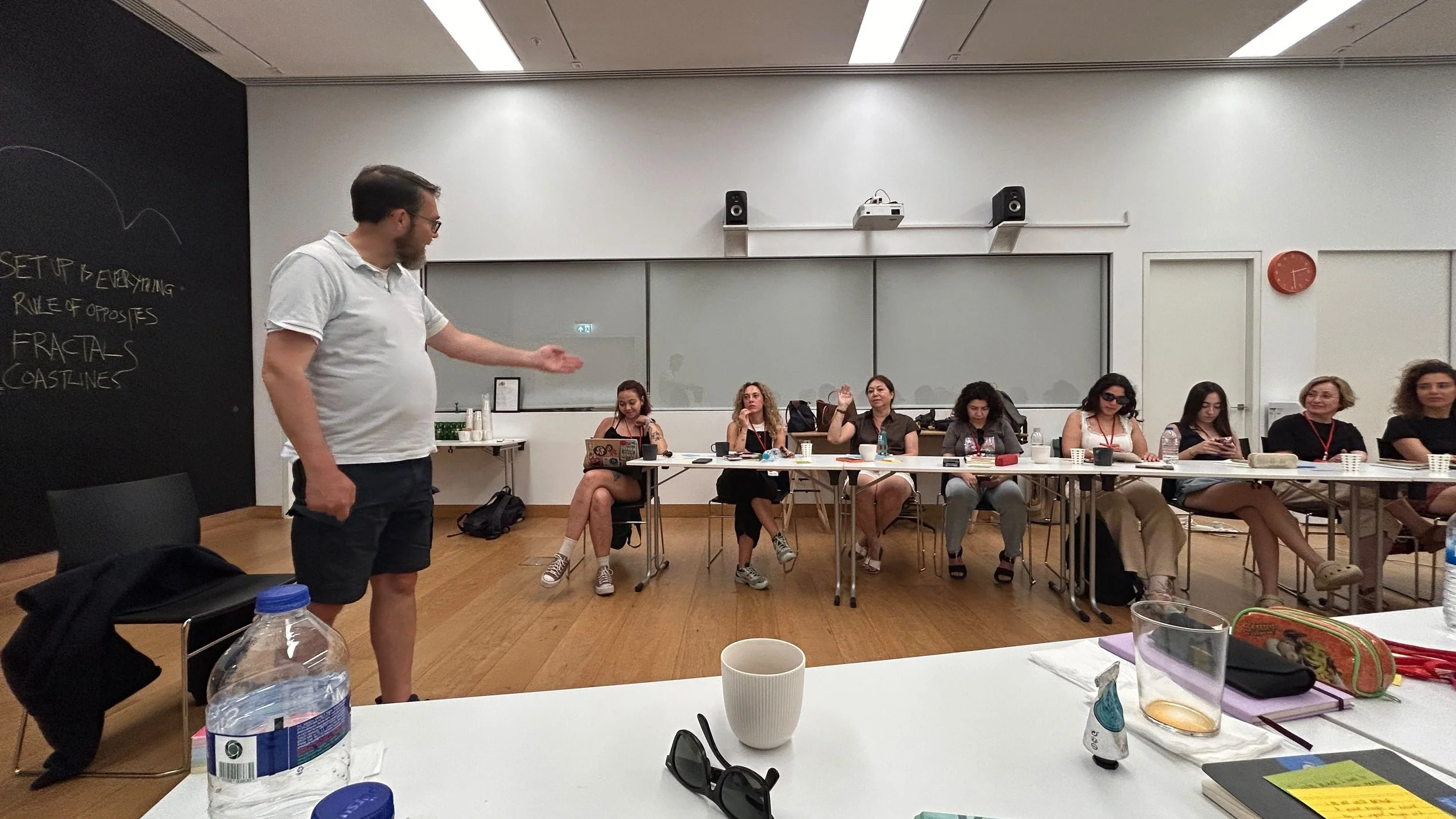 A man in a white shirt and glasses stands in front of a seated audience in a conference room, gesturing with his right hand. The audience, mostly women, are sitting at a long table with notebooks, drinks, and electronic devices, listening and some ra