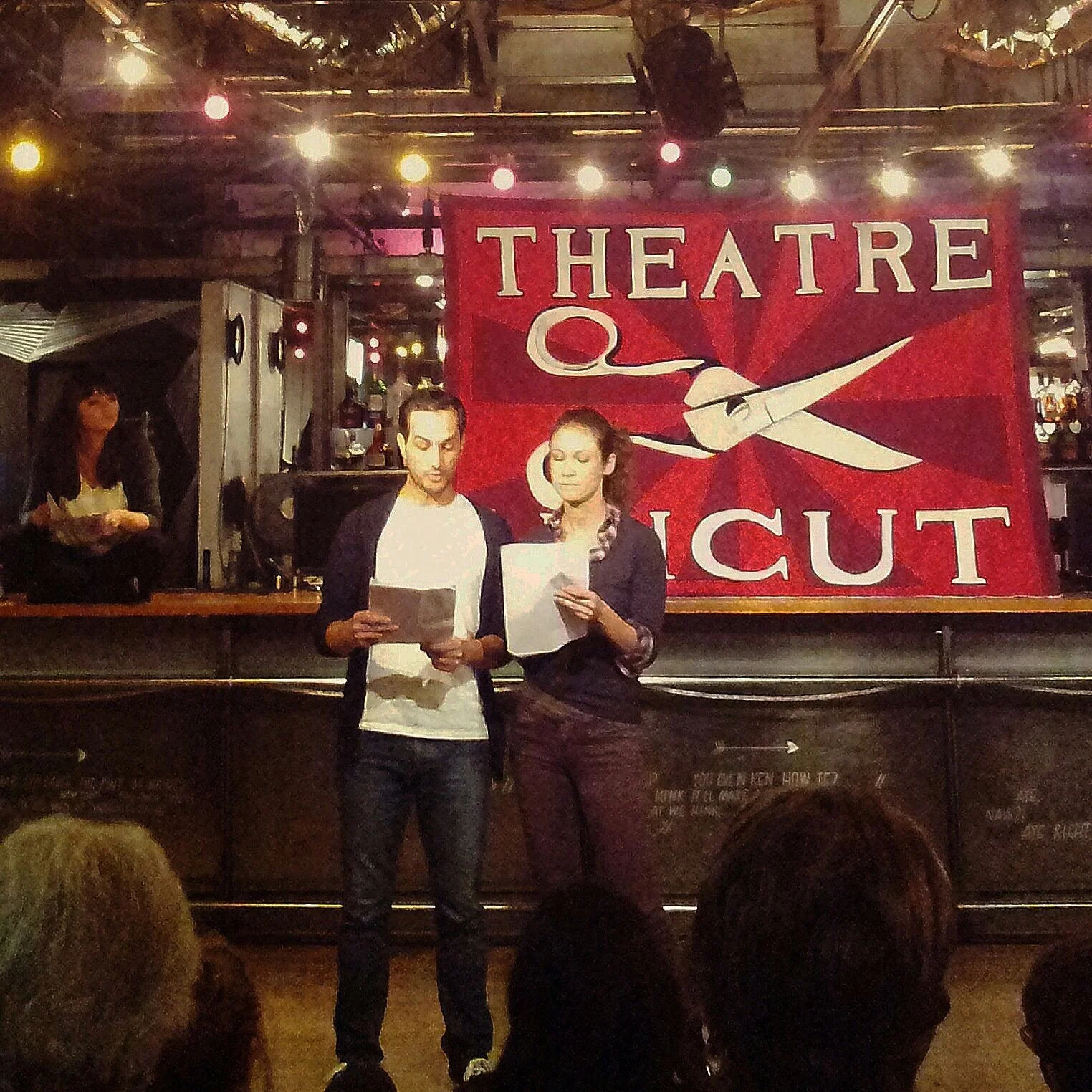 Two women standing in front of a theater poster, reading scripts, inside a venue decorated for a performance with a banner that reads 'Theatre Q' and 'Cut' featuring a graphic of a pair of scissors. Several audience members can be seen seated in fron