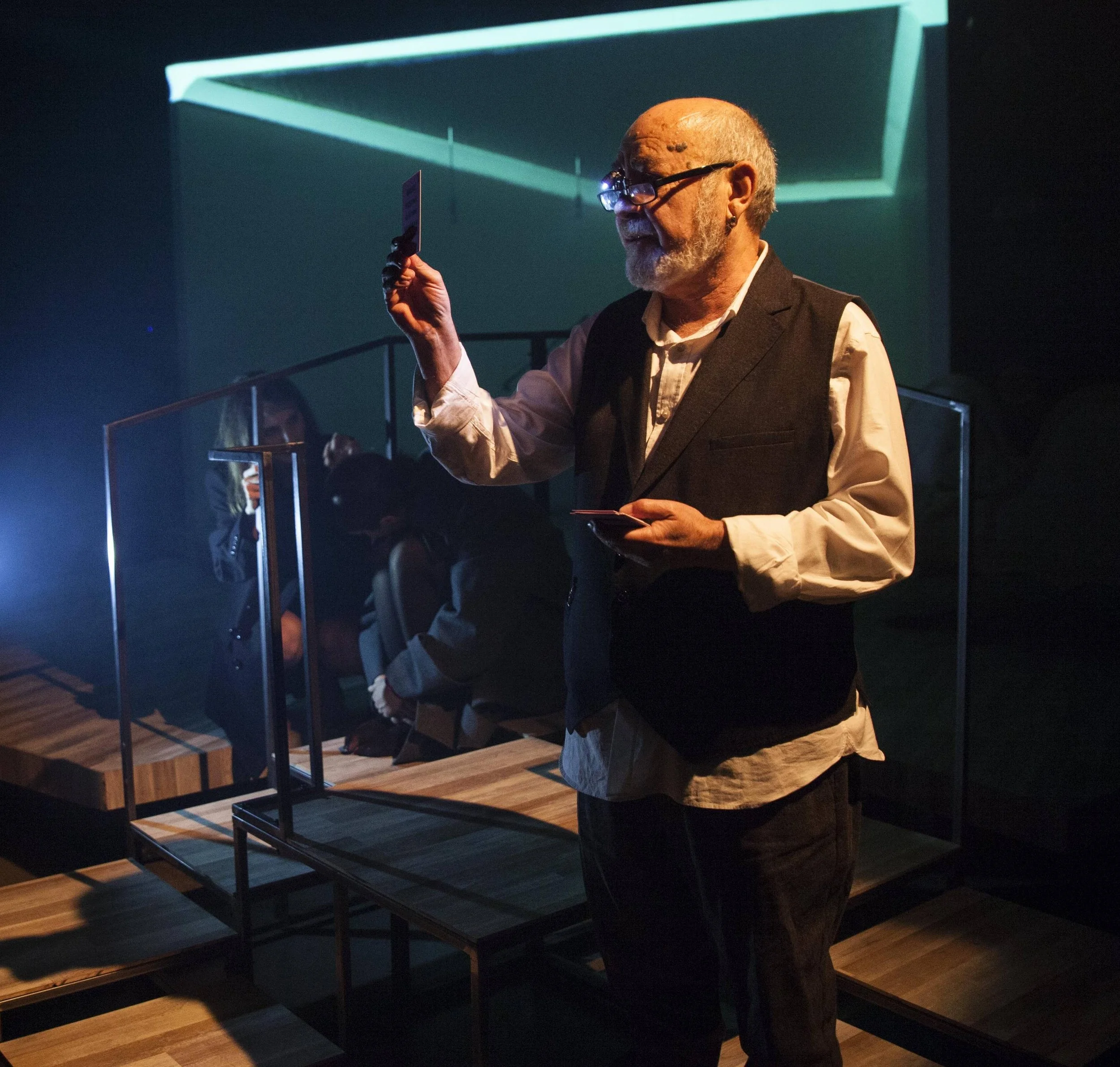 An older man with glasses, a beard, and a white shirt under a dark vest is holding a small item in his right hand and looking at it. He is indoors with a dark background and blue accent lighting, near a black metal and wood display stand.