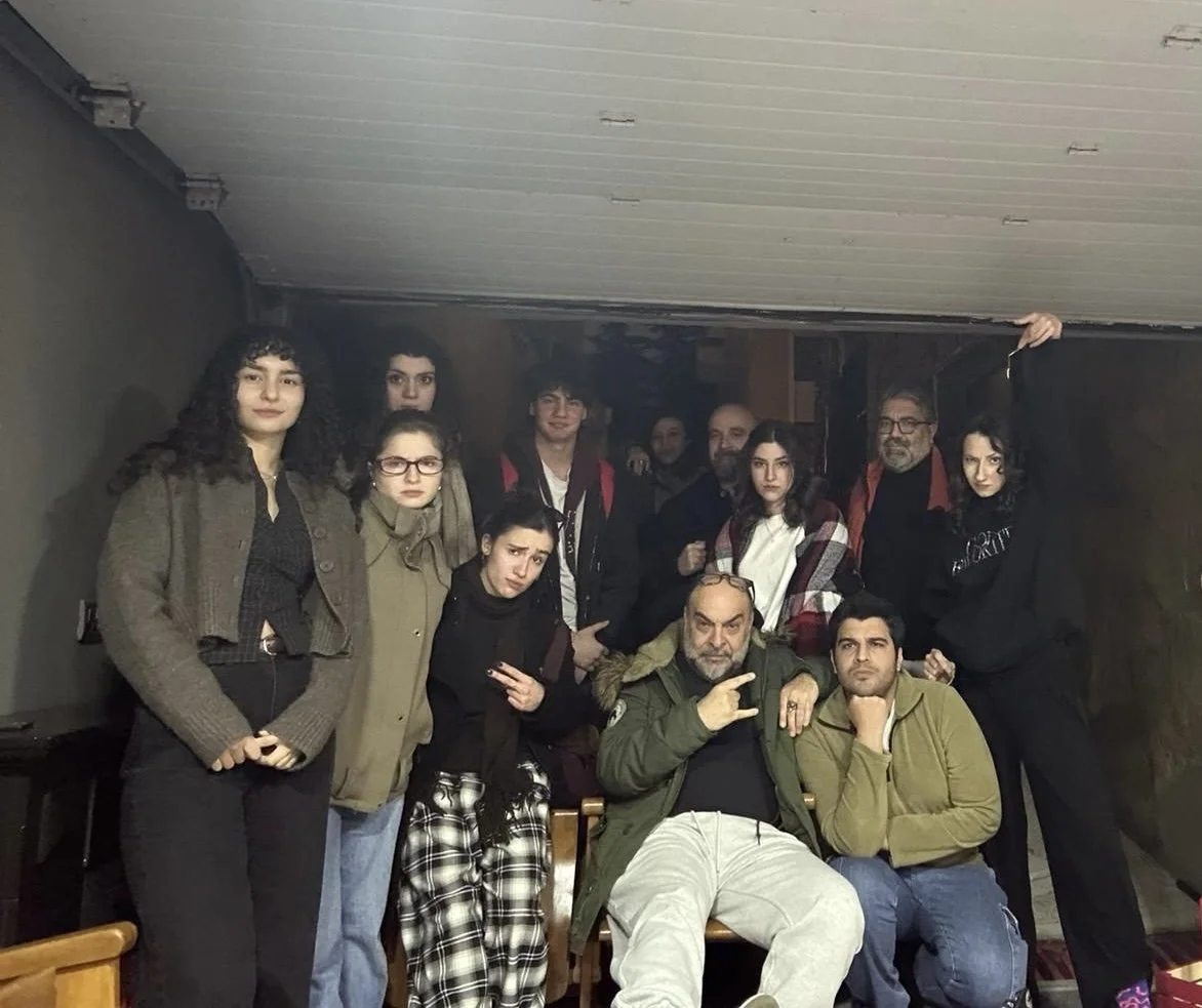 Group of people posing indoors, some sitting and some standing, with casual clothing and expressions.