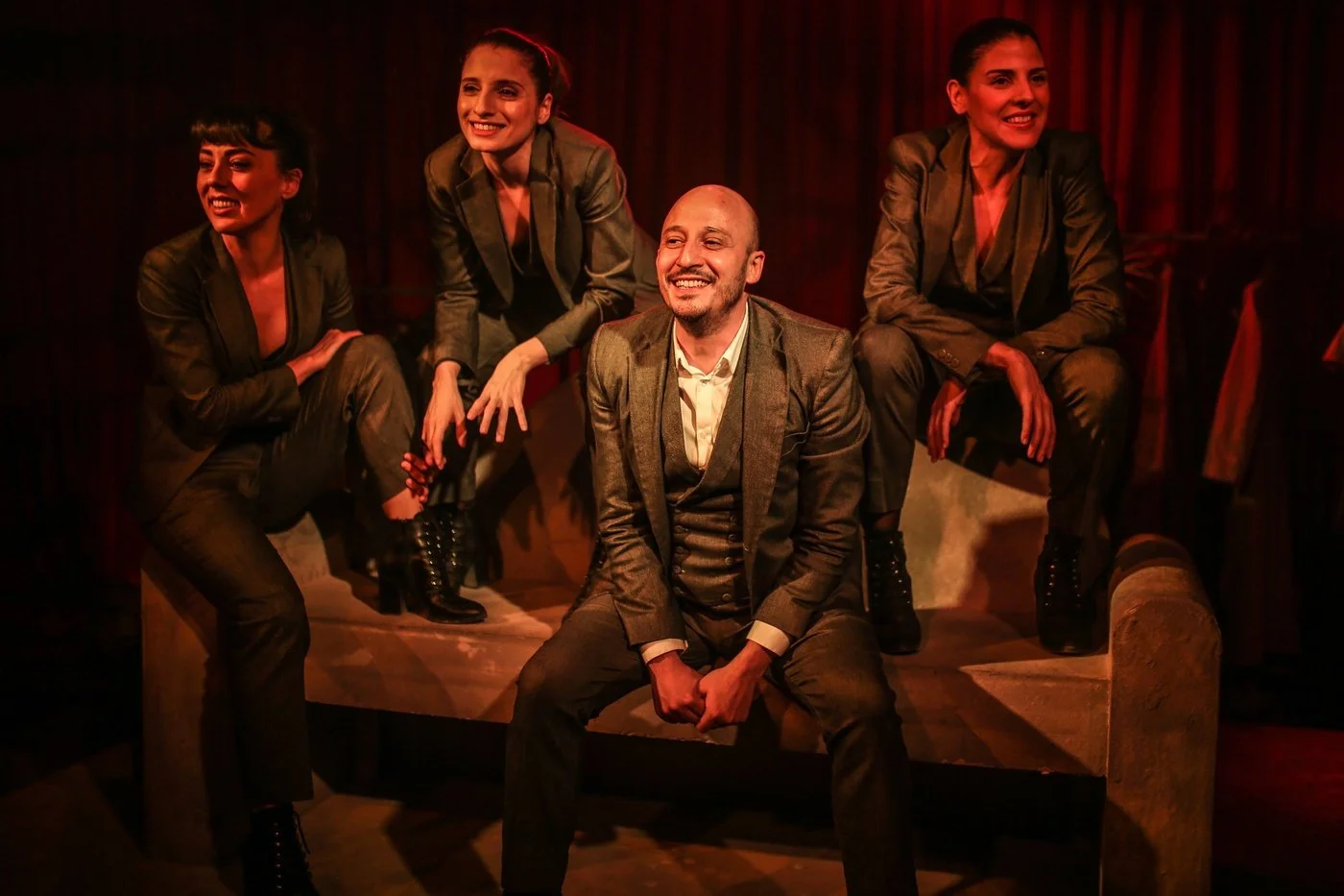 Group of four women and one man in formal suits sitting and standing on a bench, smiling, with a dark red curtain in background, possibly on a stage.