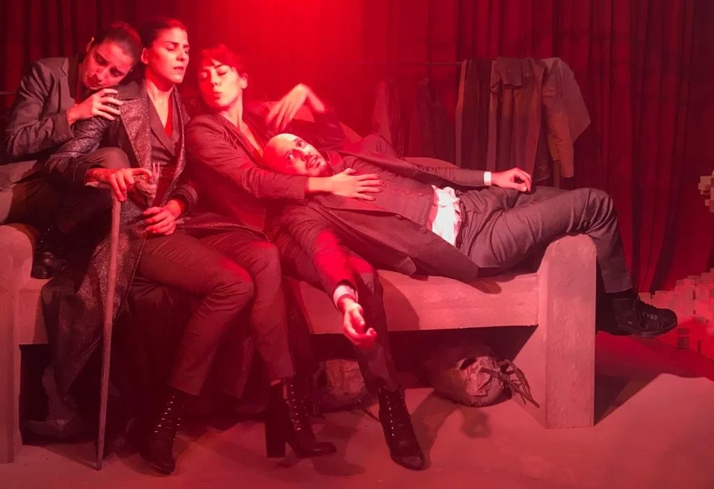 Four women sitting and lying on a stage, dressed in dark clothing, with red stage lighting and clothing hanging on a rack in the background.