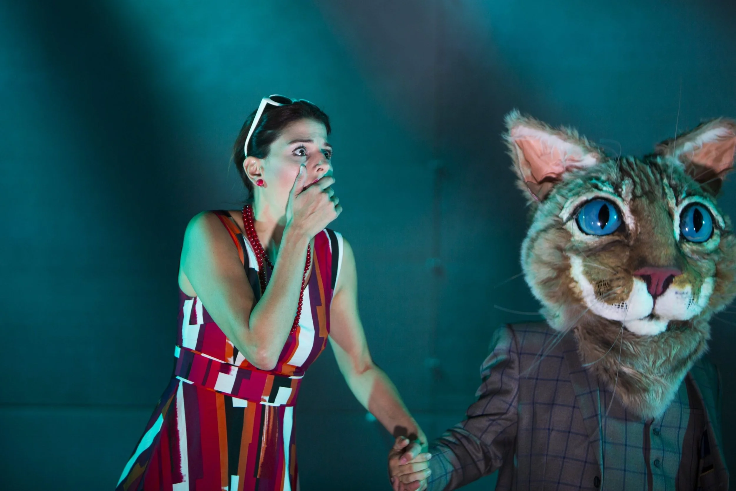 A woman in a colorful striped dress with glasses on her head covers her mouth with her hand, and a person in a large cat costume holds her hand.