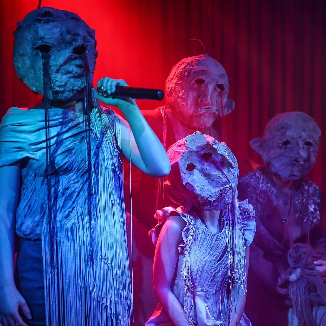 Four individuals wearing masks made of papier-mâché or similar material, with elongated features and masks resembling human faces, are standing against a red curtain at a performance or art exhibit. One person is holding a microphone.