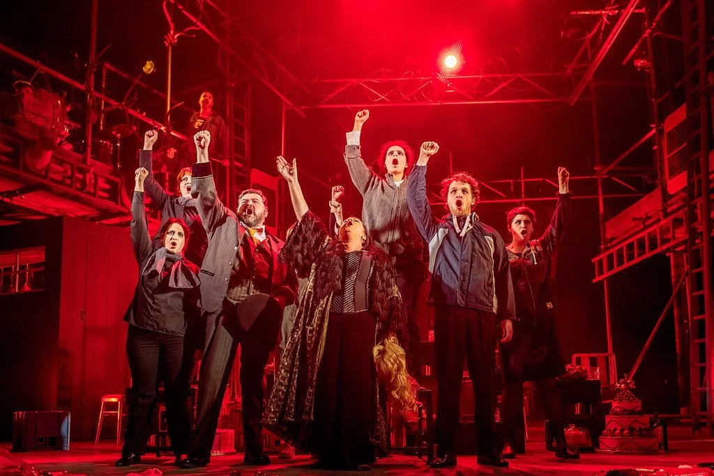The cast of a theatrical play on stage, raising their fists in a protest or anthem pose, with red lighting and stage set.