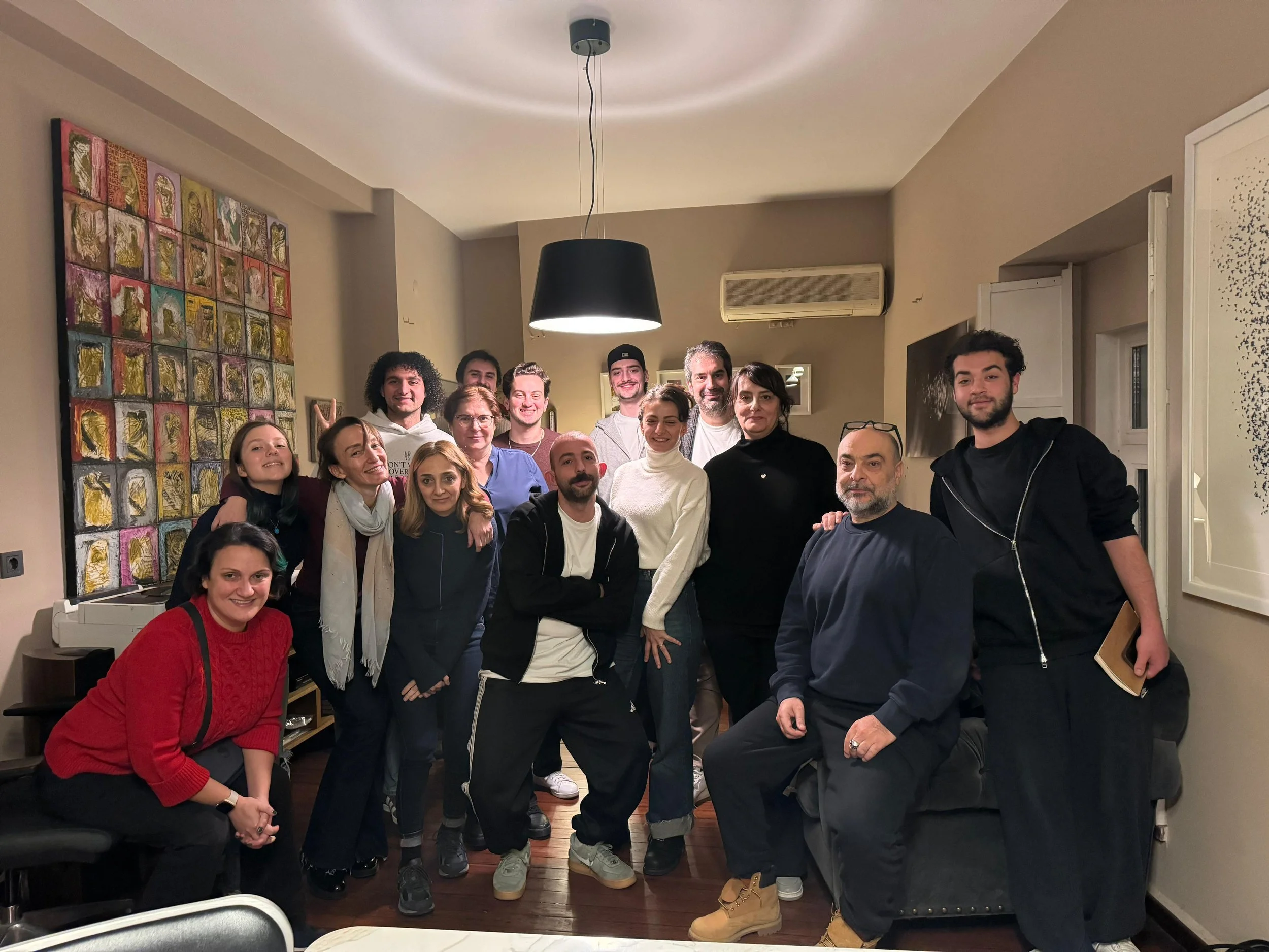 A diverse group of people gathered indoors, posing for a group photo with some smiling and others with neutral expressions. The room features a colorful abstract painting on the left wall, a ceiling light fixture, and a window with white trim on the 