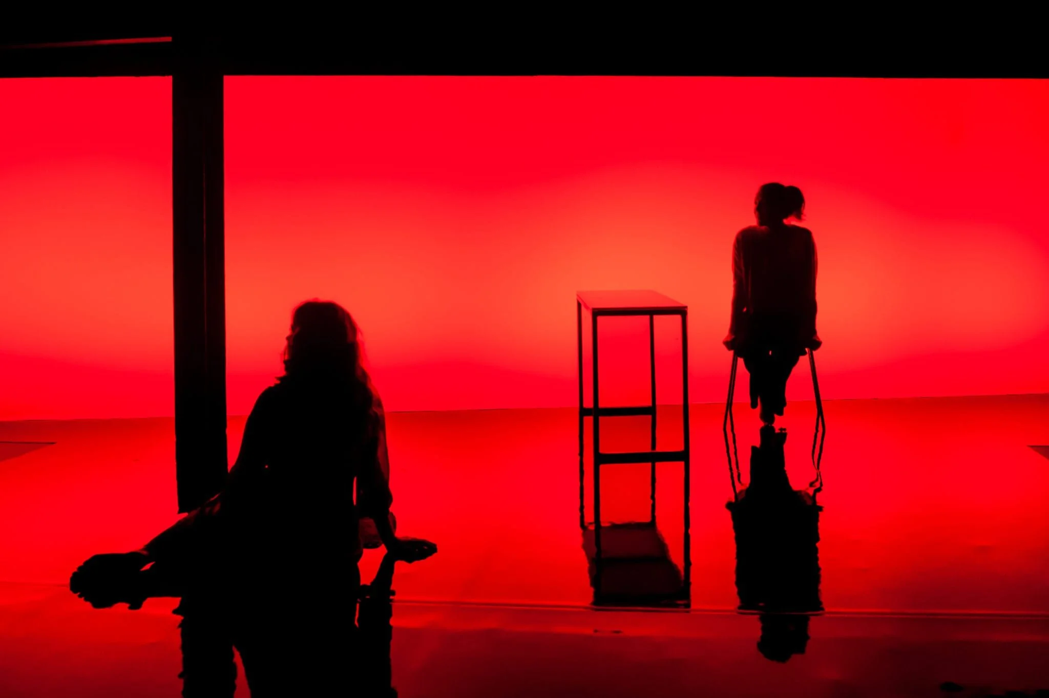 Silhouettes of three individuals in a room with red background, one sitting on the floor, one standing with crutches, and one standing behind a small table.