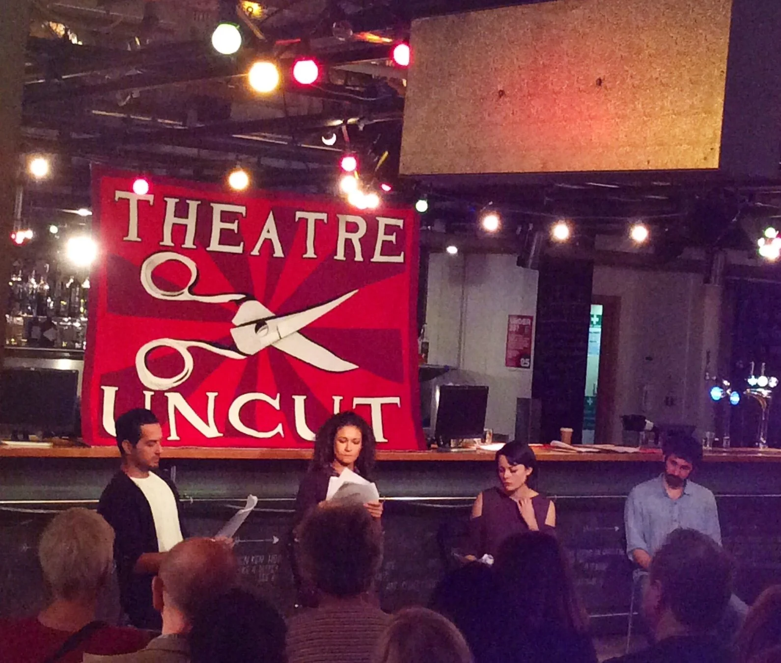 People performing at a stage with a large red banner reading "Theatre of Uncut" depicting a pair of scissors and a red star in the background, inside a venue with colorful lighting.