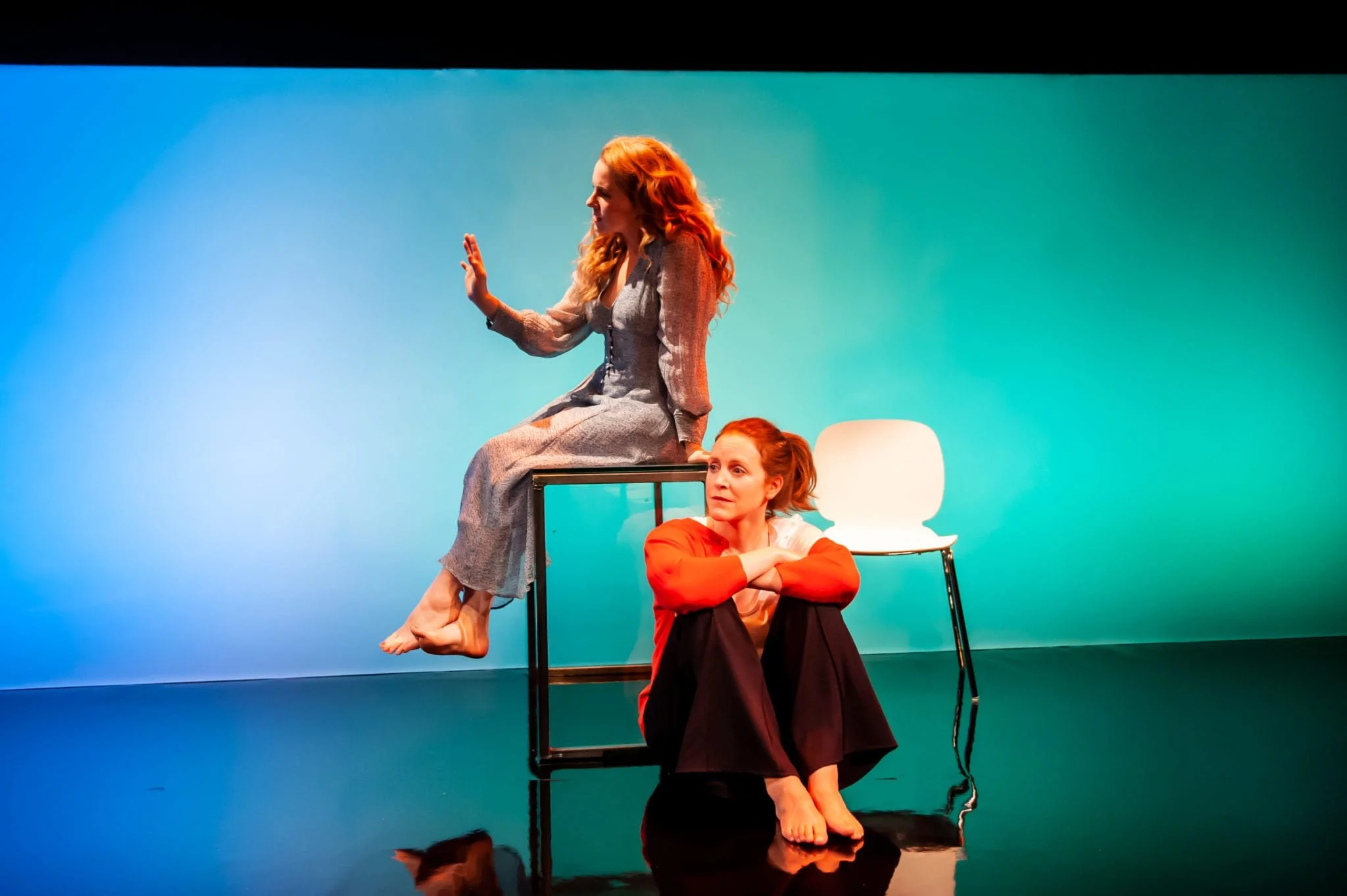Two women performing an act with one sitting on a raised surface and the other seated on the floor at a theater or studio with a teal background.