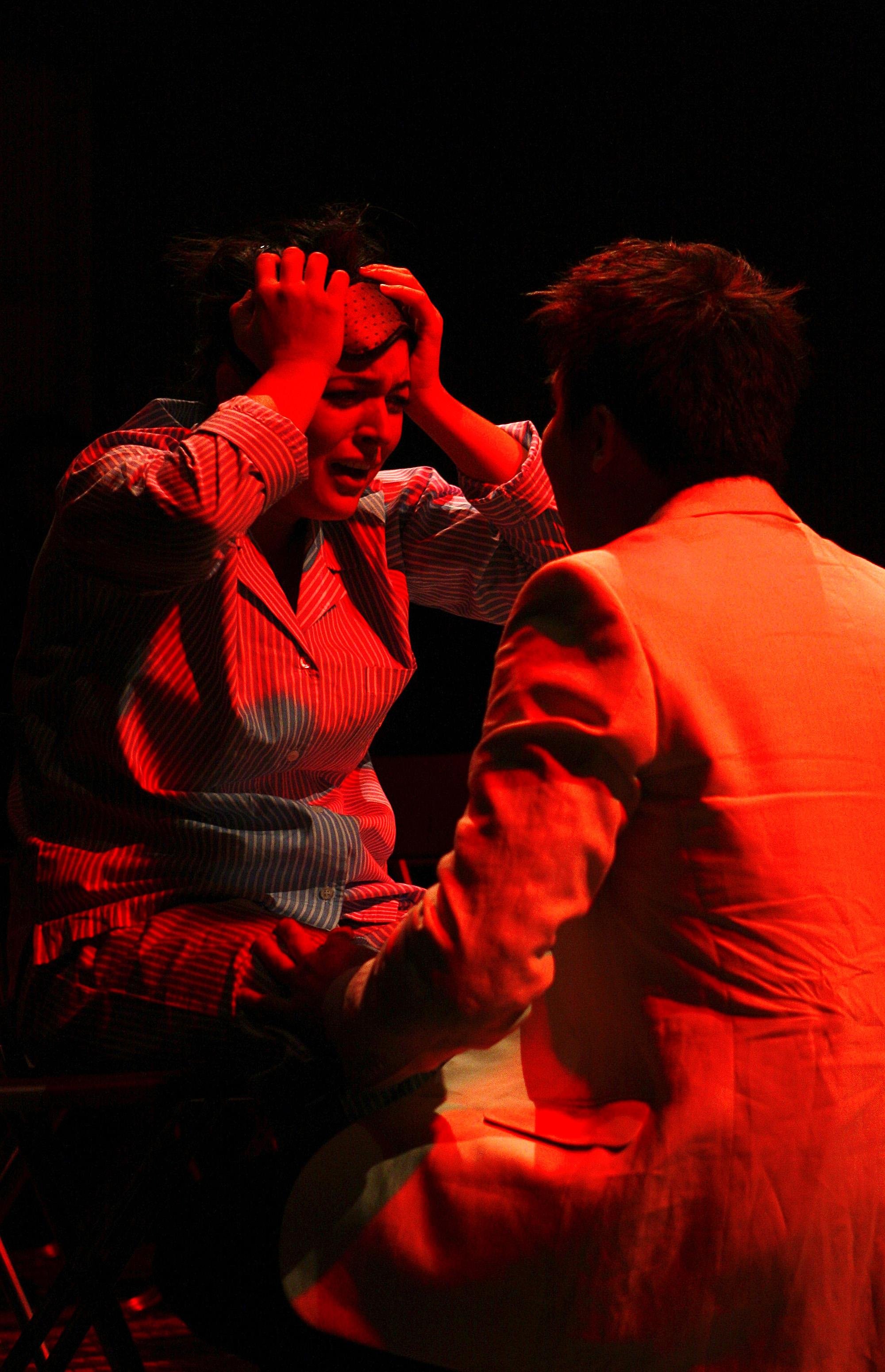 Woman in distress holding her head, sitting with another person in a red-lit environment.