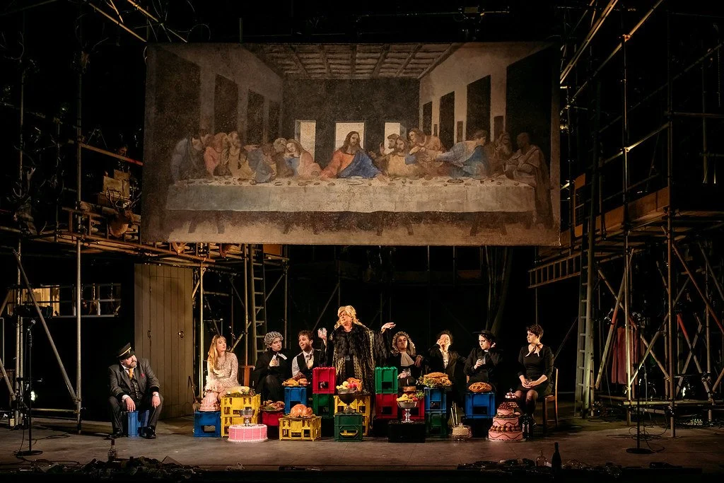 A stage scene from a theater production, with actors sitting and standing around a table with various food items and crates. A large overhead projection displays Leonardo da Vinci's "The Last Supper." The set includes scaffolding and lighting equipme