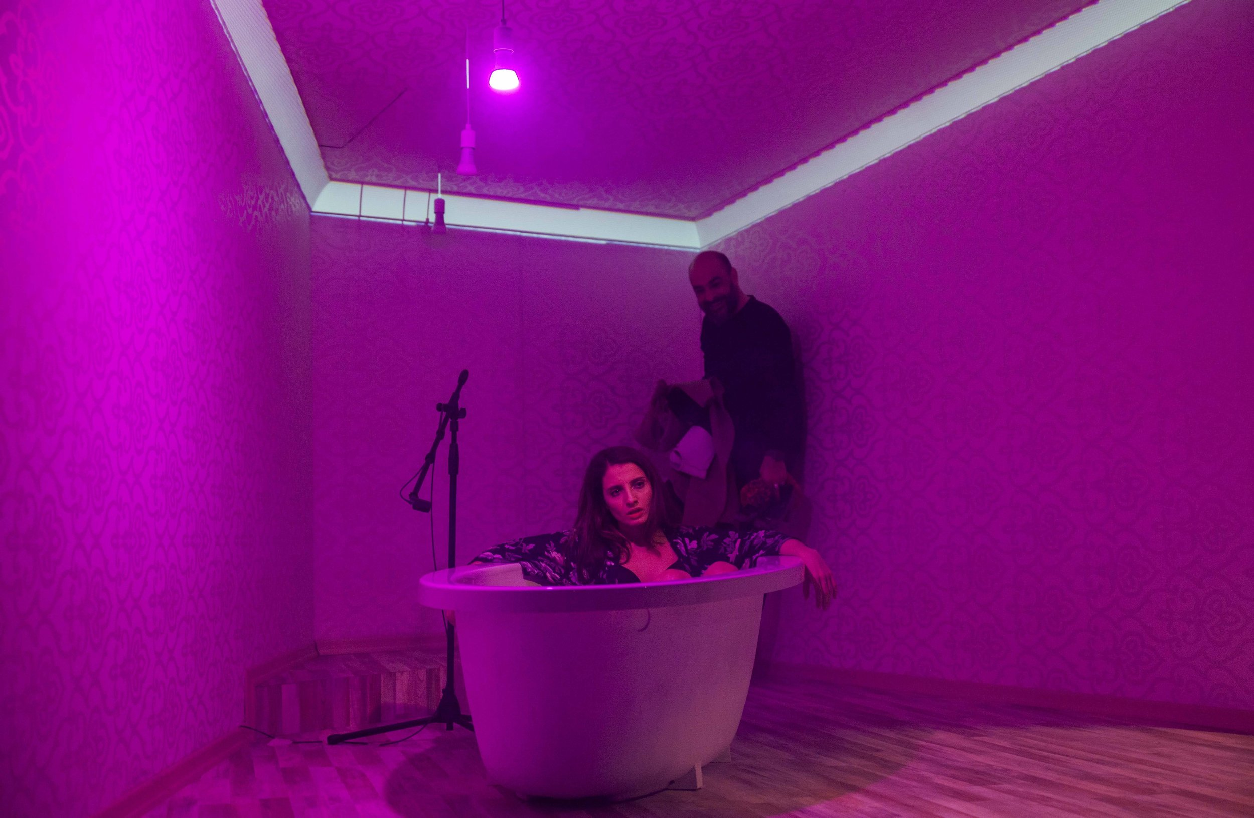 A woman with long hair sitting in a pink-lit bathtub, appearing surprised or confused, with a man standing behind her in a room with pink lighting and patterned walls.