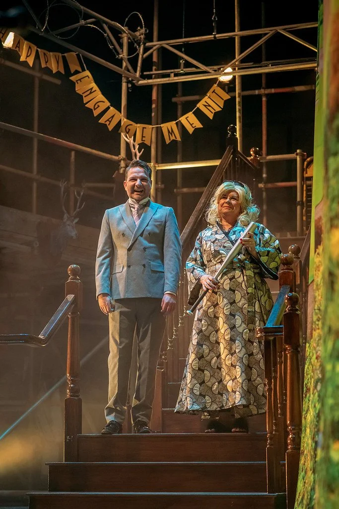 Two actors, a man in a light gray suit and a woman in a colorful patterned dress, stand on a wooden staircase on stage with a banner that reads 'Happy Engagements.' The woman holds a rolled-up document or scroll. Stage lighting and a decorated set wi