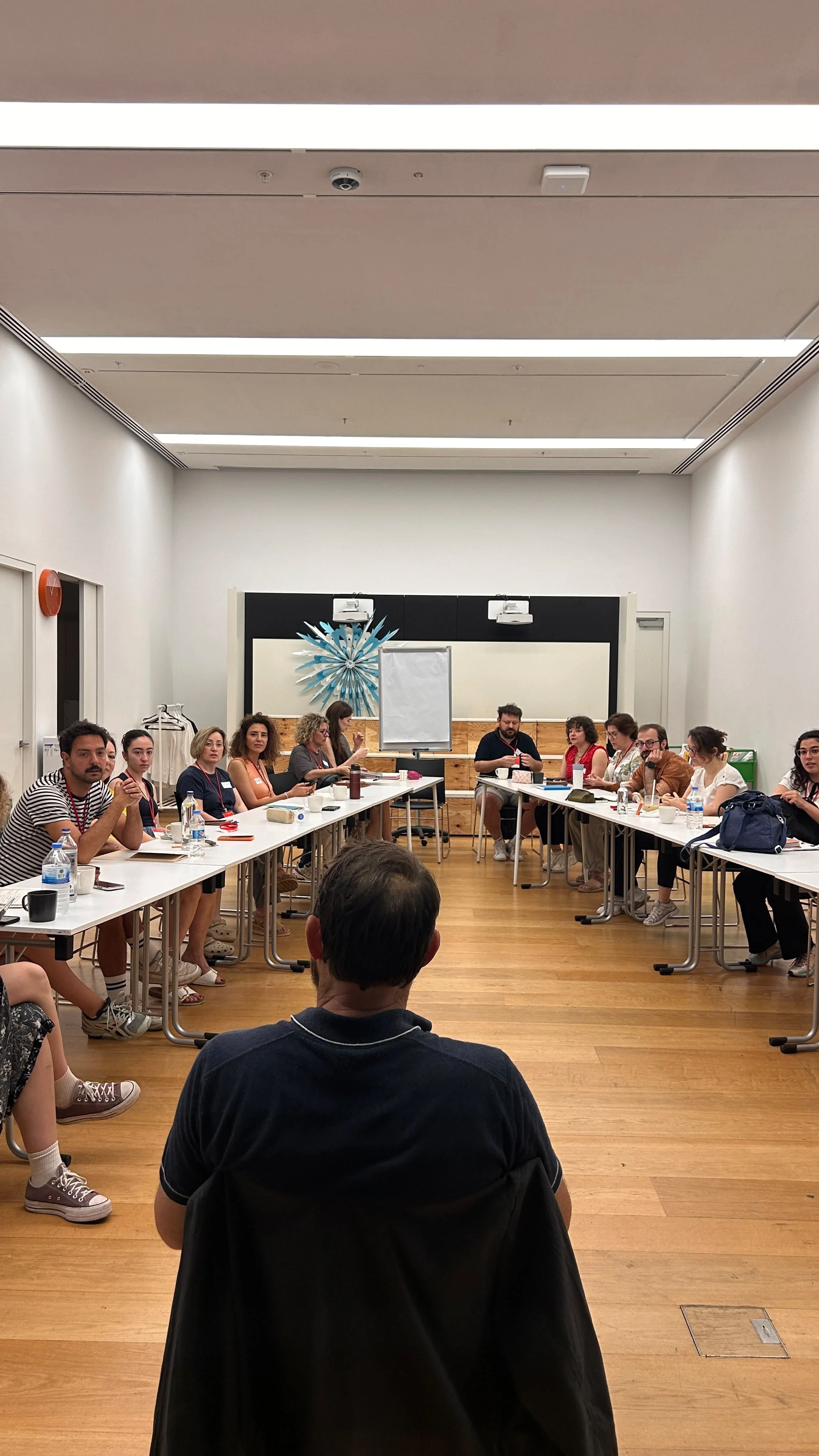 A group of people sitting around U-shaped tables in a conference room, engaging in a meeting or workshop. There is a speaker at the front near a flip chart and decorative wall art.