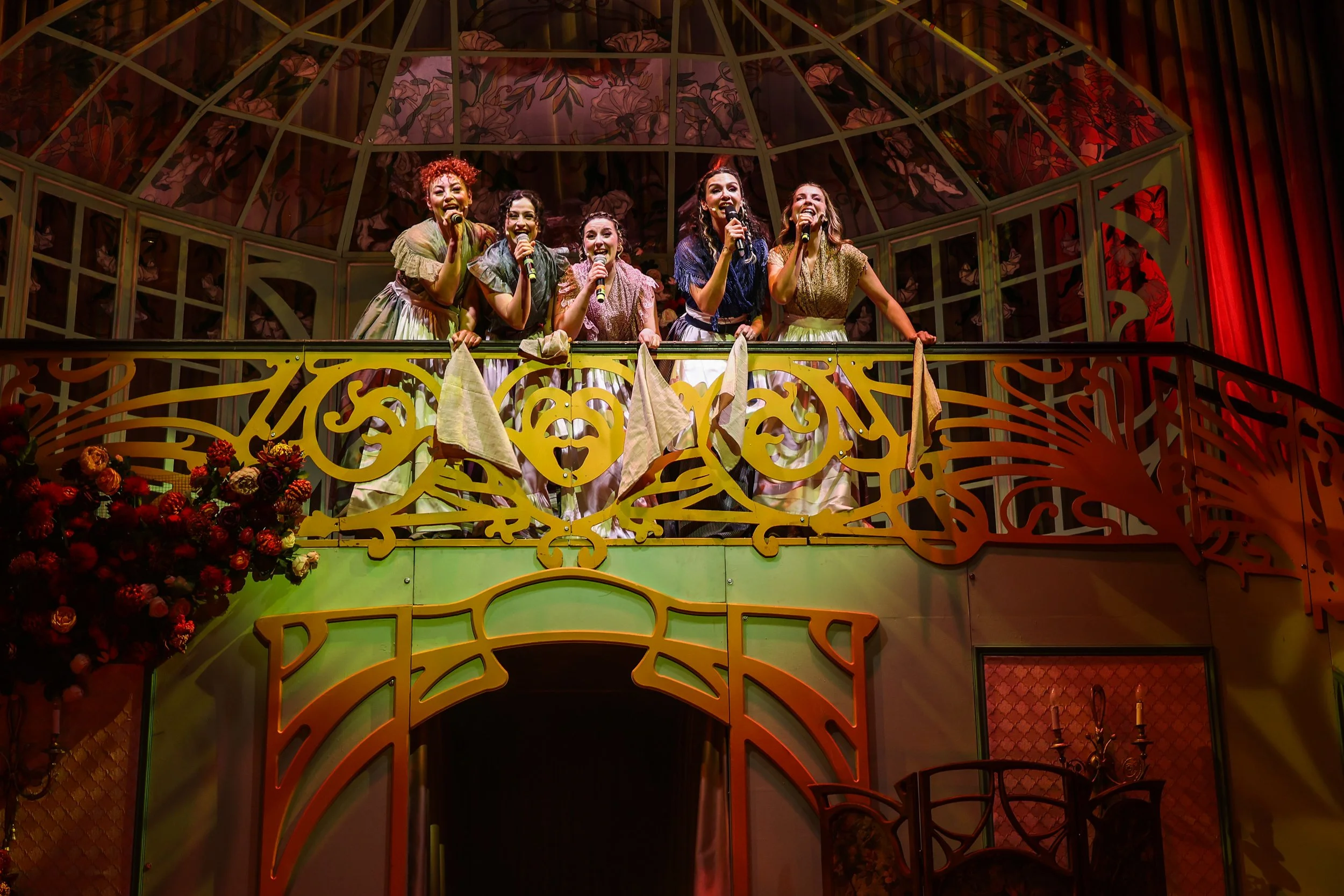 A group of five women standing on an ornate balcony wearing vintage-style dresses, holding microphones, and smiling, with a decorative floral backdrop and red curtains behind them.